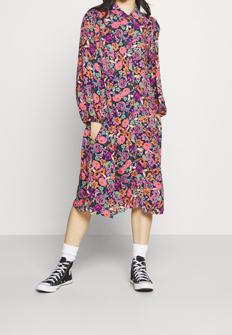 Bloemenjurk met lange mouwen, kraag en zakken. Multifunctionele bloemen op een donkere achtergrond. Zwarte high-top sneakers.