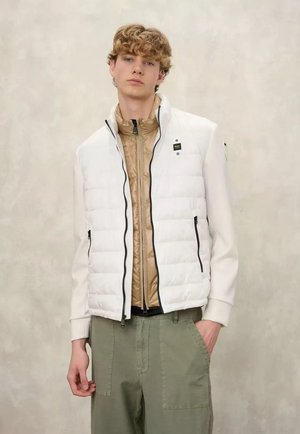 Giovane uomo con capelli ricci indossa giacche beige e bianche a strati con zip e pantaloni verde oliva, in piedi davanti a uno sfondo chiaro uniforme.