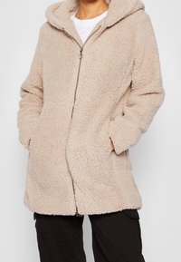 Cappotto in fleece con cappuccio beige, superficie morbida e texture, zip frontale e tasche laterali, caratterizzato da una vestibilità comoda e un design rilassato.