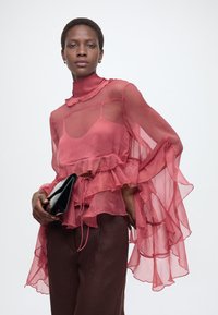 Γυναίκα που φοράει διάφανη ροζ μπλούζα με βολάν και σκούρο παντελόνι κρατάει μαύρο τσαντάκι, κοιτάζοντας με αυτοπεποίθηση την κάμερα σε άσπρο φόντο.