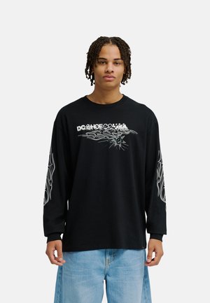 Jeune homme aux cheveux tressés portant un t-shirt noir à manches longues avec des motifs de flammes et un jean bleu, debout devant un fond blanc uni.