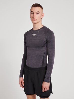 Jonge man staand, gekleed in een aansluitend grijs sportshirt met lange mouwen en losse zwarte sportshorts tegen een effen achtergrond.