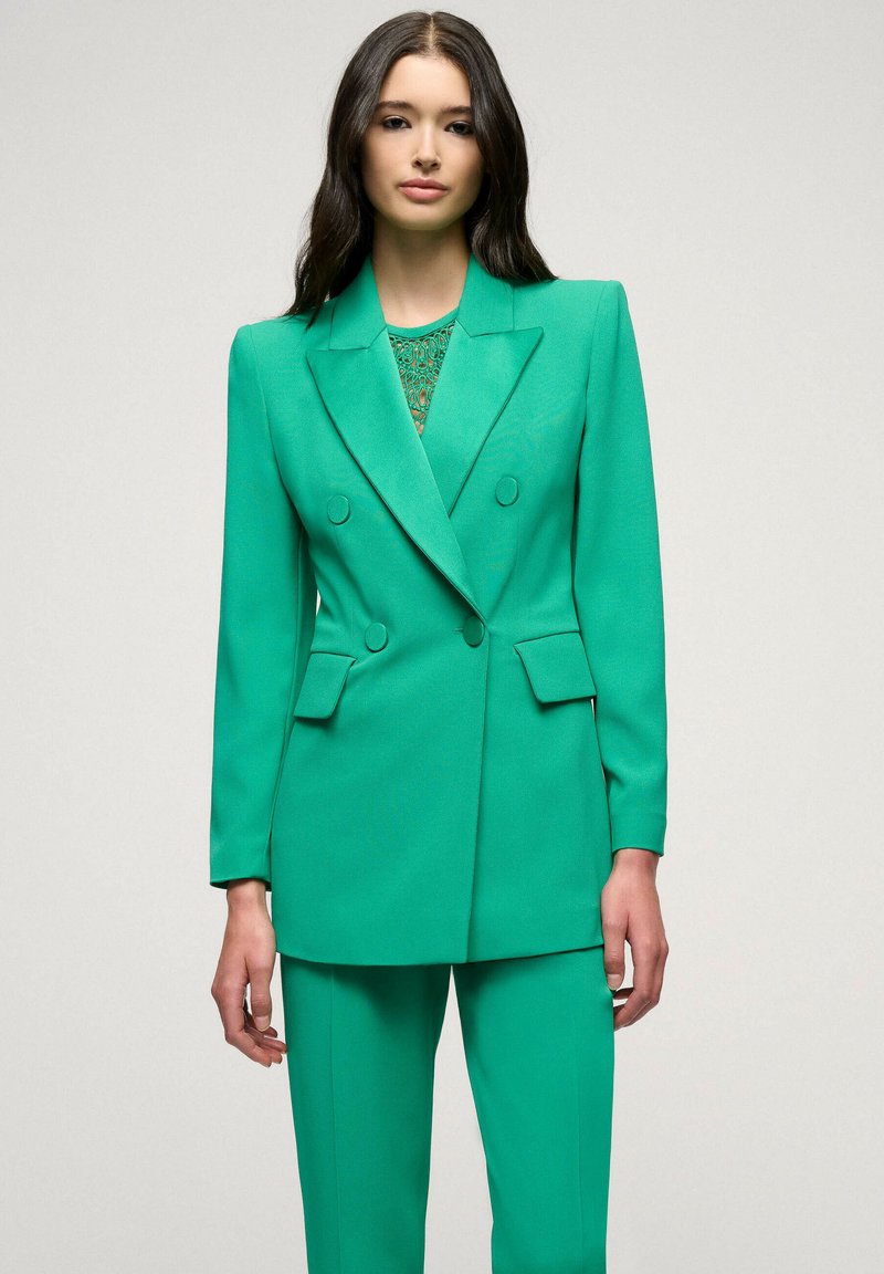 Groene op maat gemaakte blazer met dubbele knoopsluiting, aangeknipte revers en bijpassende broek. Gladde, gestructureerde stof met een slank pasvorm.