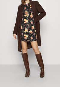 Nainen, jolla on päällään musta kukallinen mekko, tummanruskeat polven yli ulottuvat saappaat ja siihen sopiva pitkä takki, seisoo käsi lantiolla.