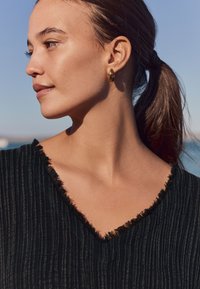 Črna zgornja oblačila z teksturo in V-izrezom s resicami. Model nosi zlat obročasto uhan. V ozadju je modro nebo in ocean.