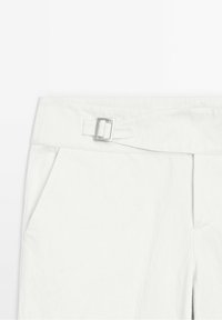 Pantalon blanc en gros plan montrant la taille avec une boucle rectangulaire argentée et une poche latérale sur fond uni.