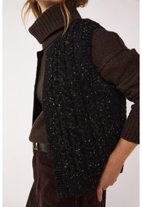 Gilet nero lavorato a maglia con motivo a treccia e apertura frontale, indossato sopra un maglione a collo alto marrone. Presenta una finitura testurizzata e nessun tipo di chiusura.