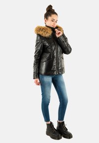 Veste en cuir noir avec col en fausse fourrure, fermeture éclair et poches latérales. Le modèle porte un jean bleu slim et des bottes noires à lacets.