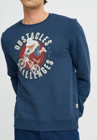 Mornarsko plava dukserica s okruglim grafičkim dizajnom biciklista i natpisom "Prepreke su izazovi." Mekani materijal, rebrasti izrez i manžeti.