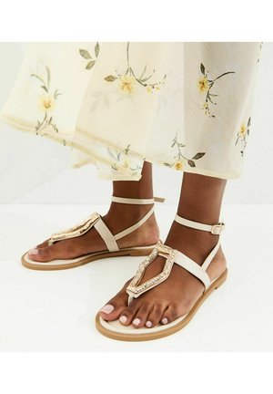 Donkere voeten met beige sandalen met decoratieve gespen, enkelbandjes en een vloeiende crèmekleurige bloemenrok.