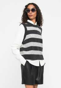 Hallhallik ja heledhallik triibuline kudumivest, mille all on valge kraega särk, koos mustade nahkšortsidega. Mudel, kellel on lokkis juuksed ja tumedad prillid.