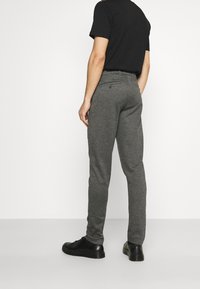 Man wearing grey textured slim-fit trousers, black t-shirt, and black leather shoes, standing against a plain light background.