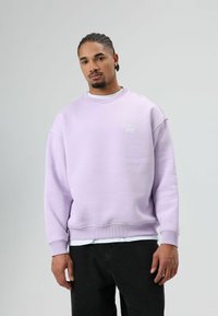 Sweat-shirt lavande avec poignets et ourlet côtelés. Présente un logo blanc imprimé sur la poitrine. Porté sur un t-shirt blanc et associé à un pantalon noir.