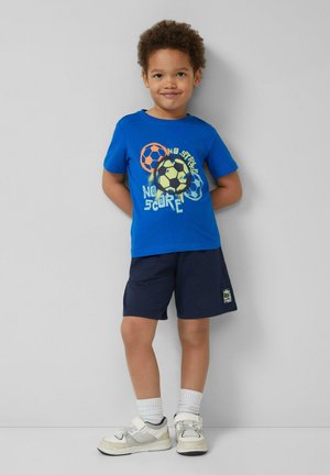 Junger Junge lächelt, trägt ein blaues T-Shirt mit bunten Fußbällen und Text, marineblaue Shorts, weiße Socken und beige Sneakers, steht an einer Wand.