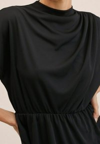 Vestido negro con una tela suave y drapeada. Presenta un escote alto y una cintura fruncida para una silueta relajada. Diseño elegante y versátil.