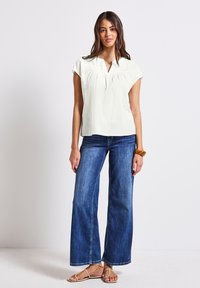 Blusa verde claro de manga corta, con pliegues horizontales, combinada con jeans de mezclilla azul de pierna ancha y sandalias de tiras color marrón claro.