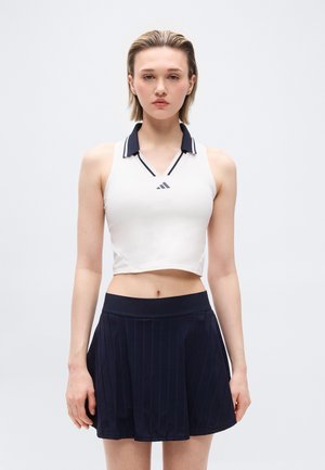 Jeune femme portant un haut court blanc sans manches avec un col noir et une jupe plissée bleu marine, debout devant un fond clair uni.