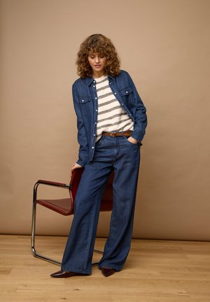 Kvinde med krøllet hår iført en cowboyjakke og vidde jeans, hvidt stribet skjorte, brunt bælte og spidse sko, stående ved siden af en rød stol.