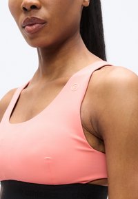 Αθλητικό κορυφαίο σε ανοιχτό ροζ με φαρδιές τιράντες, με τετράγωνο ντεκολτέ και υφασμένη μαύρη ταινία στο κάτω μέρος με λεπτομέρεια του ονόματος της μάρκας.