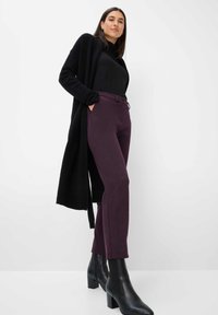 Schwarzer langer Cardigan, schwarzes enganliegendes Oberteil und strukturierte dunkelviolette Hose. Model trägt schwarze Ankle-Boots mit blockierendem Absatz.