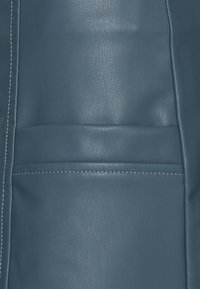 Surface en cuir bleu présentant une texture lisse, une légère brillance et des coutures blanches le long du bord ; comprend un détail de poche horizontale.