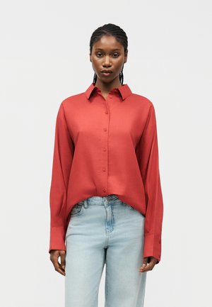 Γυναίκα με πλεξούδες, φοράει κόκκινο πουκάμισο με μακριά μανίκια και ανοιχτόχρωμα μπλε τζιν, στέκεται μπροστά σε απλό λευκό φόντο.