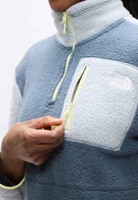 Φλις πλεκτό με φερμουάρ μέχρι τη μέση σε ανοιχτό και σκούρο μπλε, με τσέπη στο στήθος και φερμουάρ. Διαθέτει ψηλό γιακά και αντίθετες λεπτομέρειες φερμουάρ σε νέον κίτρινο.