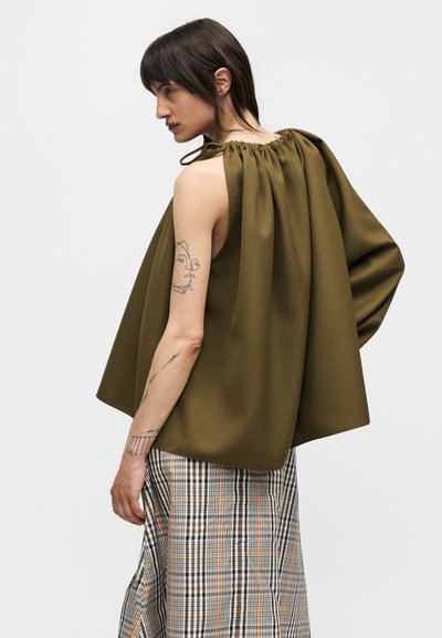 Donna con capelli scuri indossa un top senza maniche verde oliva arricciato e una gonna a quadri, mostra tatuaggi sul braccio sinistro, guarda a sinistra con un'espressione neutra.