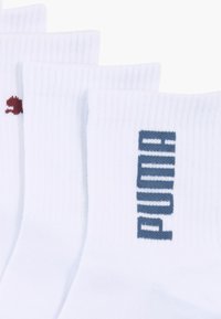 Chaussettes en coton blanches avec une texture côtelée, portant un logo "PUMA" bleu foncé et un petit emblème rouge sur un côté.