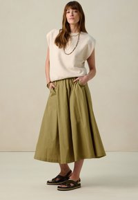 Pull à manches courtes beige, jupe verte olive à fronces avec poches latérales, et sandales noires à lanières avec un léger talon.