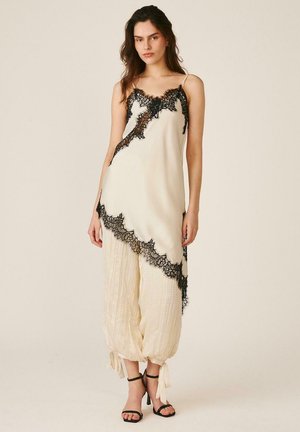 Mujer con vestido lencero color crema con ribete de encaje negro, pantalones crema sueltos recogidos en los tobillos y sandalias negras de tacón alto, de pie contra un fondo liso.