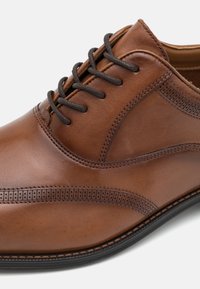 Chaussure habillée en cuir marron avec une texture lisse, présentant des coutures décoratives et des accents perforés sur le côté, et des lacets foncés.