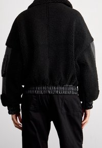 Schwarze Schaffelljacke mit hohem Kragen, elastischen Bündchen und einem elastischen Saum. Mit Lederakzenten an den Ärmeln und einer strukturierten Oberfläche.