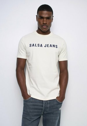 Hombre con camiseta blanca de manga corta con el texto "SALSA JEANS" y vaqueros azules, de pie con las manos en los bolsillos frente a un fondo liso.