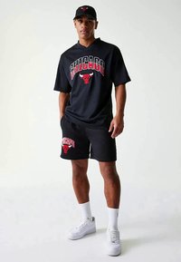 Homem a usar um boné preto dos Chicago Bulls, uma camisola preta e calções com o logo da equipa, meias brancas e ténis brancos, de pé contra um fundo branco.