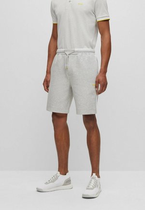 Mann trägt hellgraue Shorts mit Kordelzug, ein passendes hellgraues Polo mit gelber Einfassung und weiße Turnschuhe und steht vor einfachem Hintergrund.