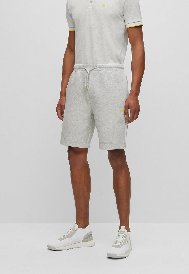 Homme portant un short gris clair à cordon, un polo gris clair assorti avec un liseré jaune, et des baskets blanches, debout sur un fond uni.