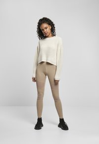 Pull cropped en tricot crème associé à des leggings beiges avec des accents en pièce. Le modèle porte des chaussures de sport noires. Fond neutre.