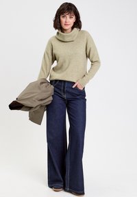 Pull en maille vert clair à col roulé, jean bleu foncé à jambe large avec des coutures visibles, et une veste grise drapée sur le bras.
