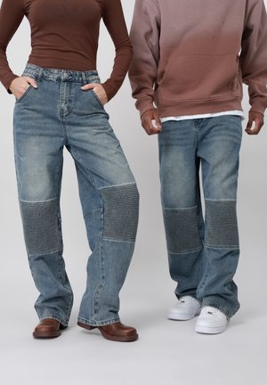 Dos personas llevan jeans azul claro con parches acolchados en las rodillas, una con botas marrones y camiseta marrón, la otra con zapatillas blancas y sudadera marrón.