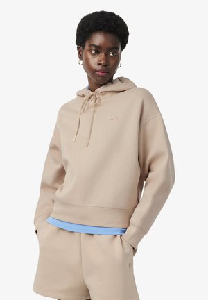 Femme portant un sweat à capuche beige et un short assorti avec un t-shirt bleu clair en dessous, les mains dans les poches, regardant vers la droite sur un fond blanc.