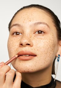 Lepestift blyant i hånden, påfører en varm brun nyanse. Huden viser lette til mørke fregner; modellen har klare brune øyne og minimal sminke.