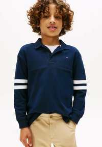 Marineblaues Langarm-Polohemd mit weißen Streifen an den Ärmeln, Knopfleiste am Kragen und kleinem Logo. Getragen mit beigen Hosen. Weiche Baumwolltextur.