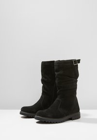 Une paire de bottes noires en daim mi-mollet avec des semelles en caoutchouc plates, une fermeture éclair sur le côté et une sangle à boucle près du haut.