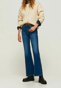 Beżowy sweter z dzianiny, niebieskie dzwony, czarne botki za kostkę. Teksturowany materiał, luźny krój i ciemny dżins o klasycznym fasonie.