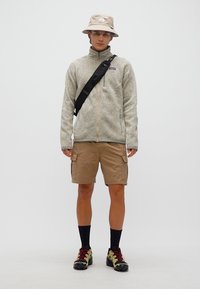 Νέος άνδρας που φοράει μπεζ καπέλο τύπου bucket, ανοιχτό γκρι φλις με φερμουάρ, καφέ cargo σορτς, μαύρη τσάντα χιαστί, μαύρες κάλτσες και παπούτσια πεζοπορίας με κόκκινα κορδόνια.