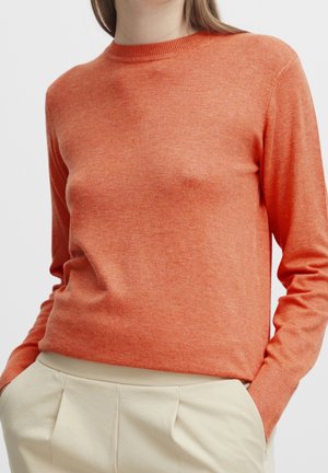 Frau trägt einen eng anliegenden orangenen Langarmpullover, in hochtaillierte cremefarbene Plisseehosen gesteckt, die Hände in den Taschen.