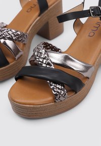 Sandalias de plataforma trenzadas en negro y metálico con tacón de bloque, que cuentan con una plantilla marrón y una correa ajustable en el tobillo.