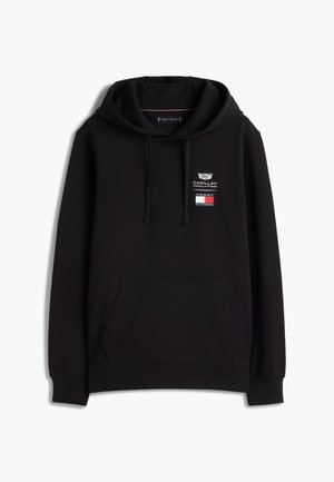 Νεαρός άνδρας φορώντας μαύρο φούτερ με κουκούλα με τα λογότυπα Cadillac και Tommy Hilfiger, άσπρο παντελόνι και λευκά αθλητικά σε λευκό φόντο.