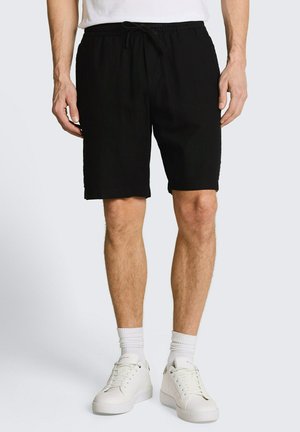 Homme portant un short à cordon noir, des chaussettes blanches, des baskets blanches et une chemise blanche, debout sur un fond clair uni.